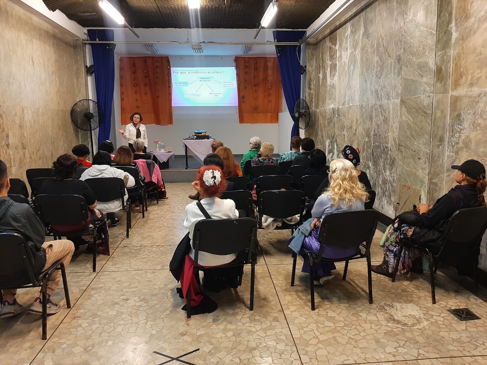 19 pessoas se sentam em cadeiras pretas, viaradas para frente. Uma mulher branca de cabelos cacheados, vestindo blusa branca e calça preta, está na frente de todos falando. Ao lado dela, sob um palco cinza, há uma mesa coberta por toalha roxa com uma jarra de água em cima e outra mesa ao lado, também coberta com toalha roxa. Em cima da mesa, há um projetor preto que projeta uma apresentação de PowerPoint na parede branca. Em volta da apresentação há uma cortina laranja. Dos dois lados do palco há ventiladores pretos e uma cortina azul.